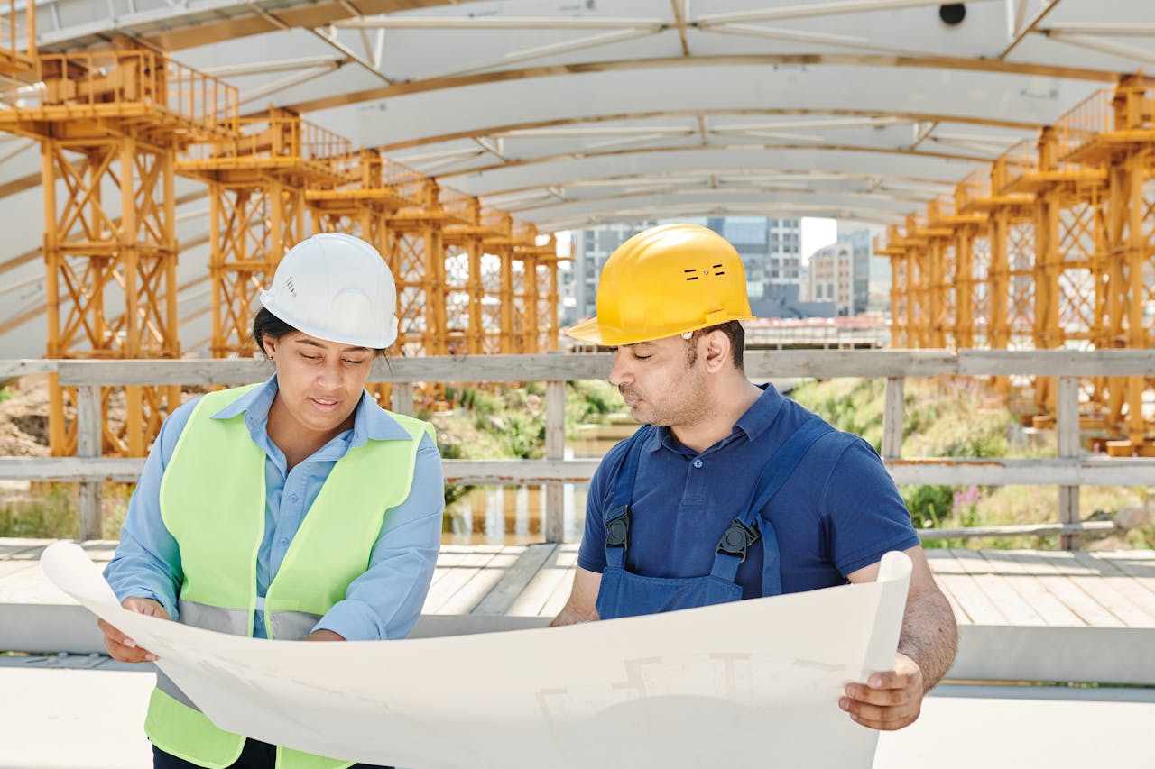 EXPERIENCIA EN DOCENCIA UNIVERSITARIA Engineering team analyzing blueprints together at an outdoor construction site with safety gear.